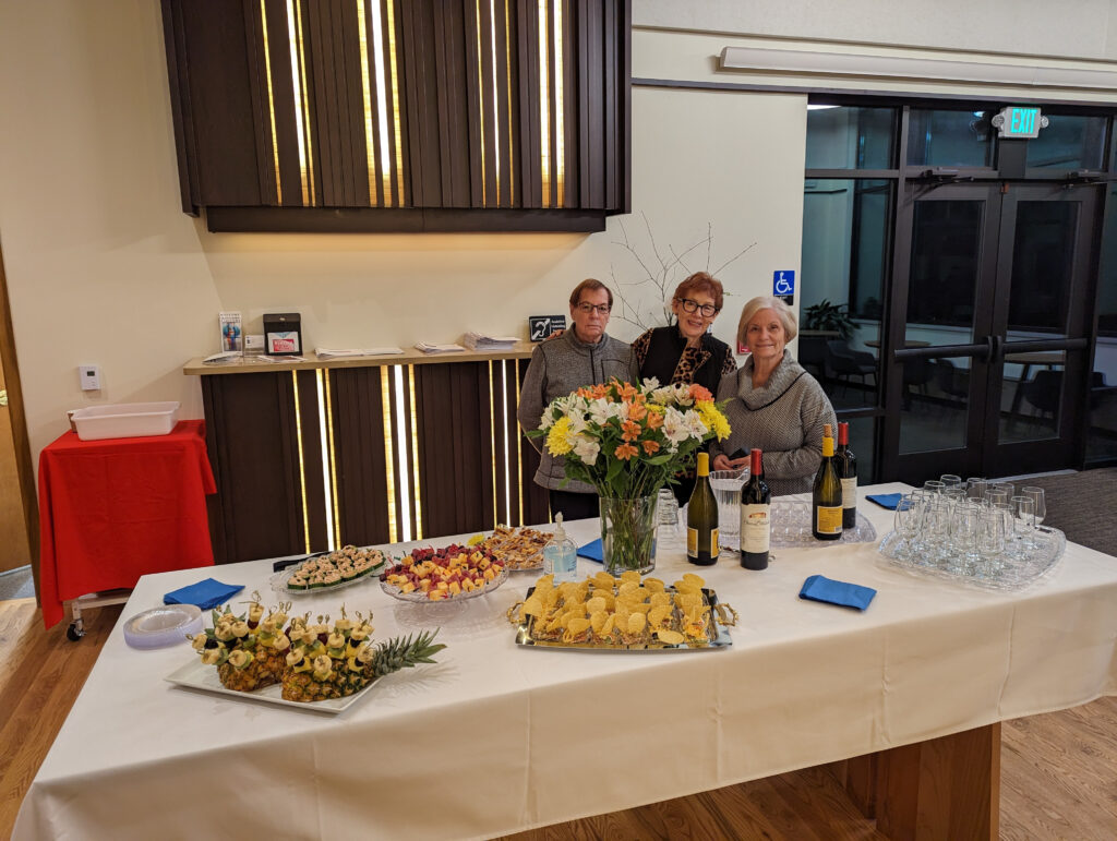 Three women standing behind a table of food and wine