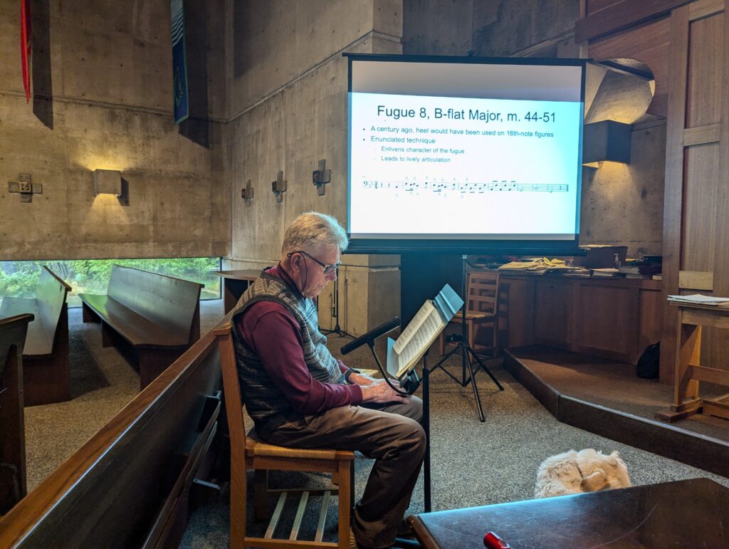 A seated man is looking at a music book. There is a screen with text on it in the background.