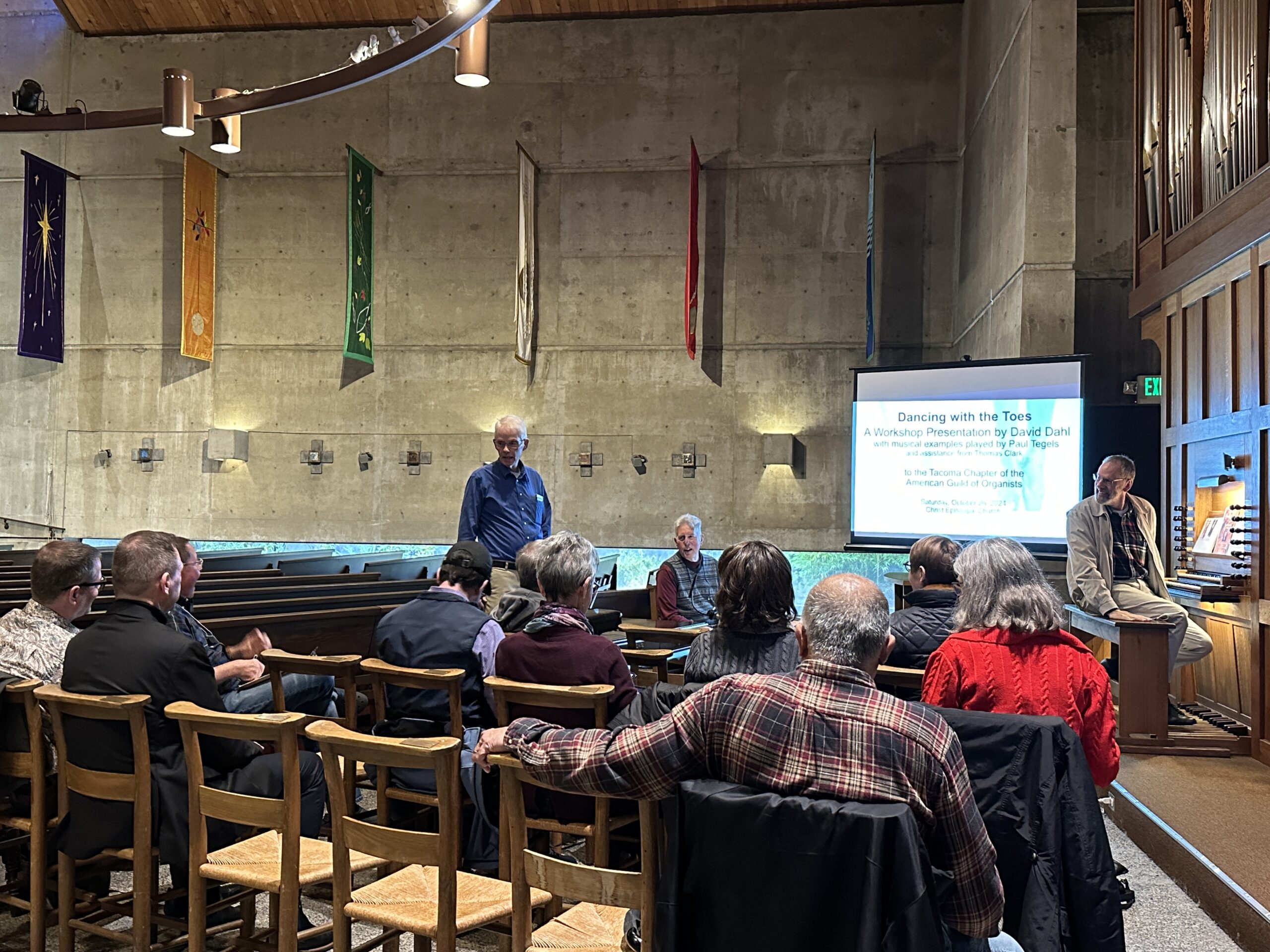 A group of people faces a screen showing text, with one person standing and talking, and another seated on the bench of an organ.