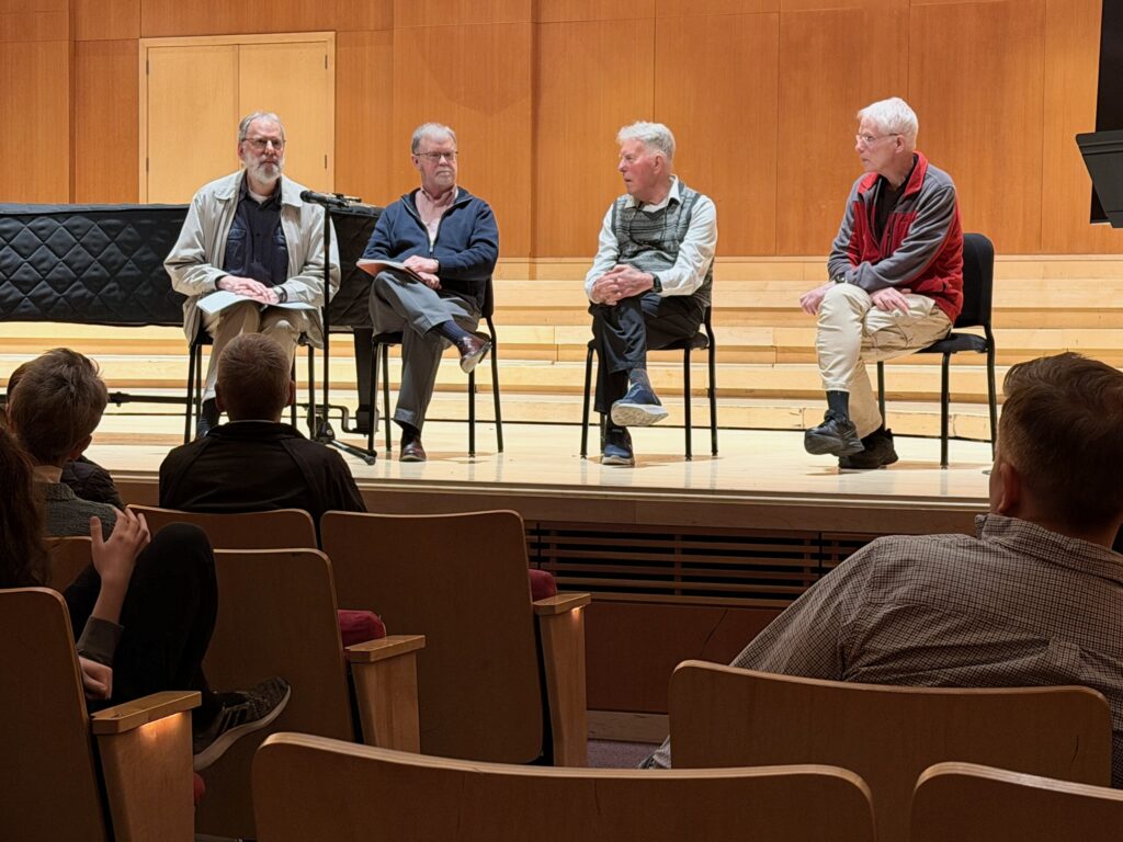 Four men sit on the stage in front of an audience